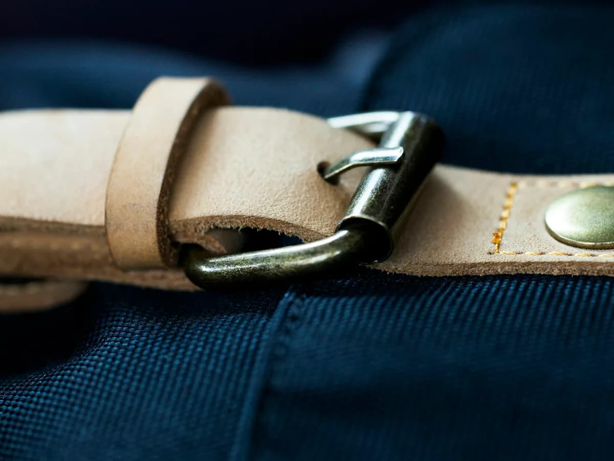 Natural tan leather belt with a metal buckle resting on dark fabric.
