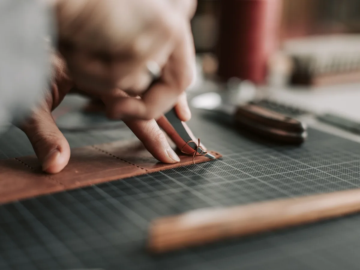 Hands cutting a brown leather piece on a gridded workshop mat.